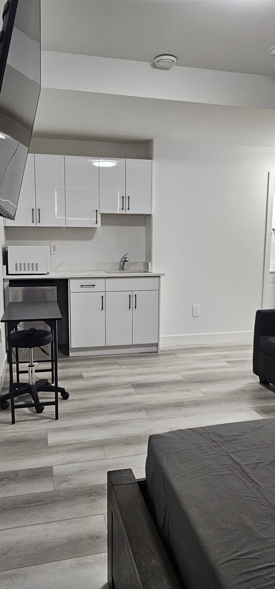 A compact kitchenette is shown, featuring white cabinets and a small sink. A black bar table is positioned nearby. The light-colored flooring enhances the modern feel of the space, with soft natural light entering from a nearby window, illuminating the overall clean design.