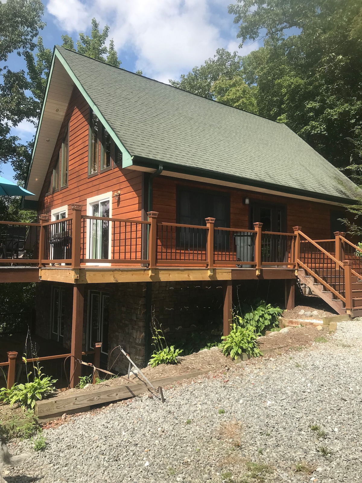 The home features a warm wood exterior complemented by a green roof. The spacious deck wraps around the structure, providing access to outdoor areas. Natural landscaping includes gravel pathways and lush greenery, enhancing the serene atmosphere of the property.