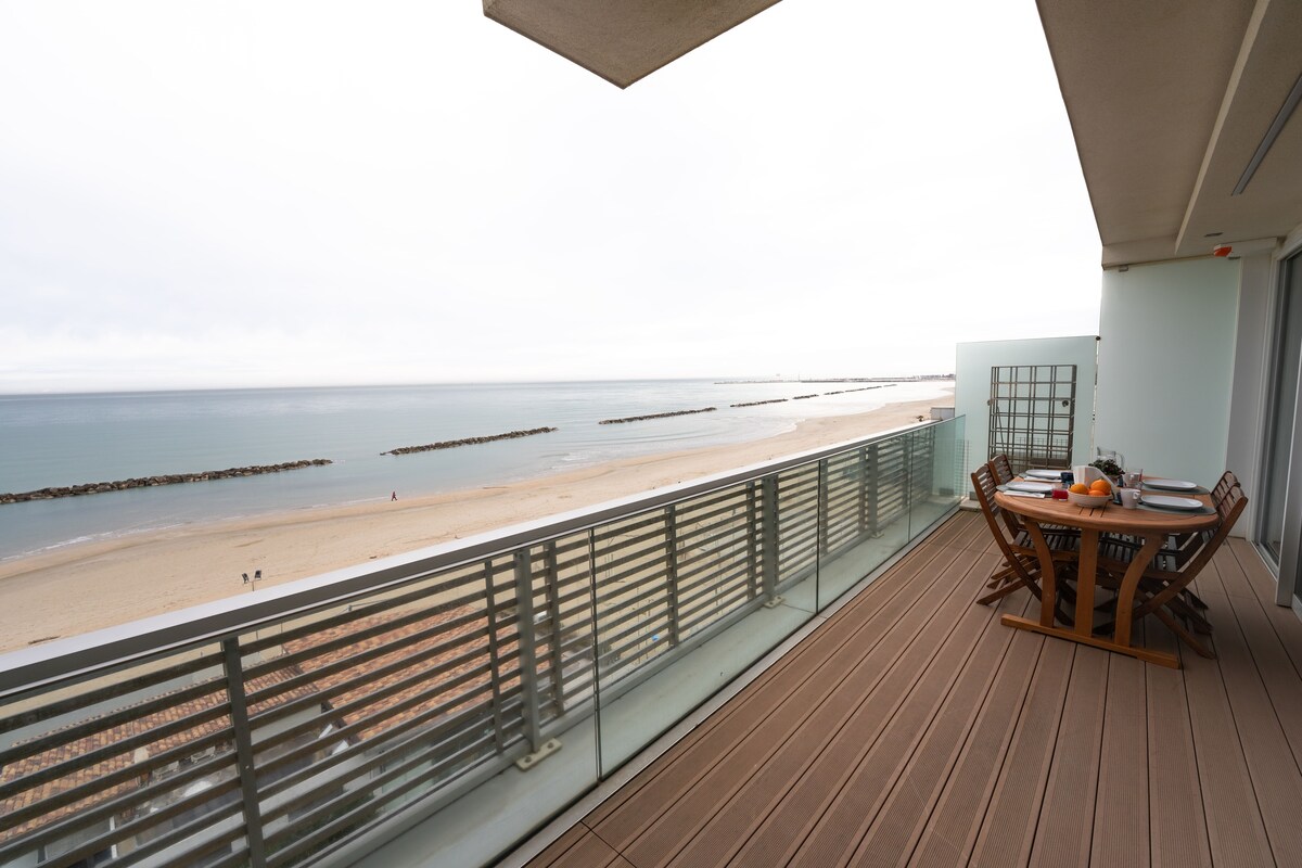 An outdoor terrace features a wooden dining table surrounded by six folding chairs, offering unobstructed views of the beach and sea. The serene shoreline stretches out in the distance, framed by a glass railing that enhances the open-air experience.