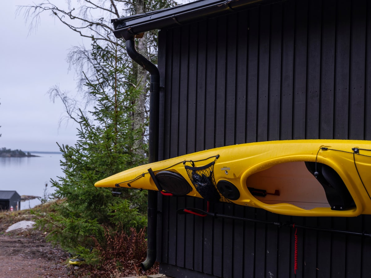 A bright yellow kayak is securely mounted on the side of a dark wooden building. The calm waters of the fjord are visible in the background, framed by a few trees, creating a serene setting for outdoor activities.