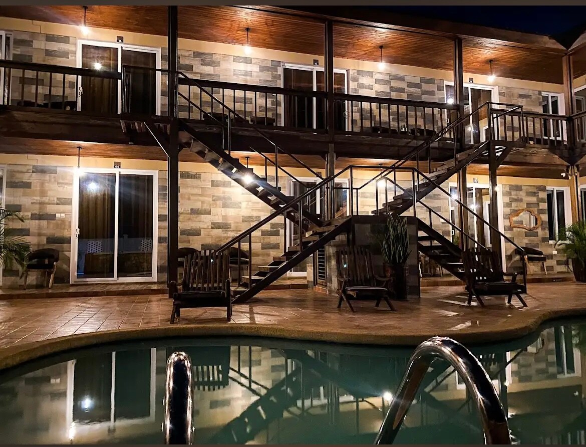 Two levels of a modern building are displayed, featuring wooden balconies and illuminated pathways. A sparkling pool reflects the structure's warm lights. Wooden lounge chairs are positioned around the pool area, creating a relaxing outdoor atmosphere.