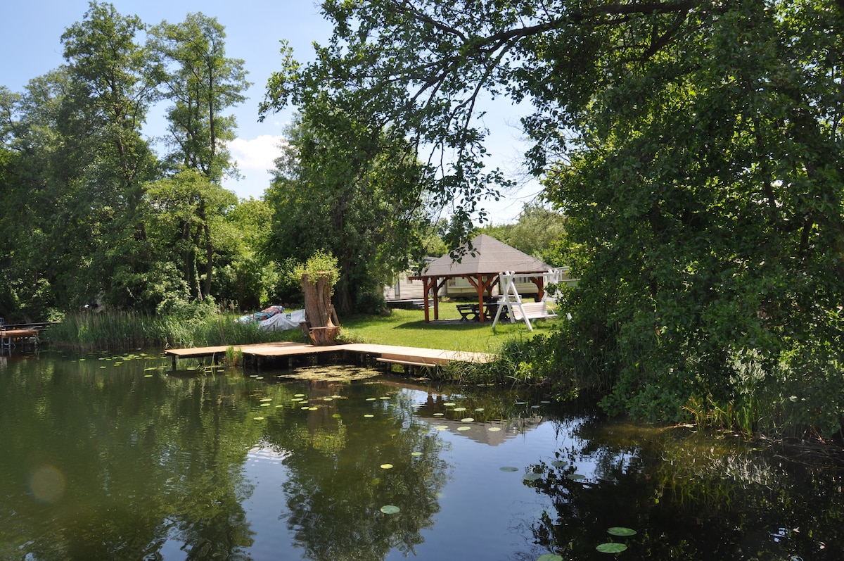 The image captures a serene lakeside scene featuring a well-maintained yard with lush greenery. A wooden gazebo is positioned nearby, complemented by a wooden dock extending into the calm water. Trees surrounding the area create a sense of privacy and tranquility.