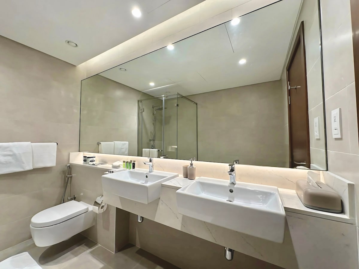 A modern bathroom features dual sinks set within a sleek countertop. A large mirror expands the space, reflecting the minimalist design. Clean lines and neutral tones create a calming environment, complemented by subtle amenities arranged neatly on the countertop.