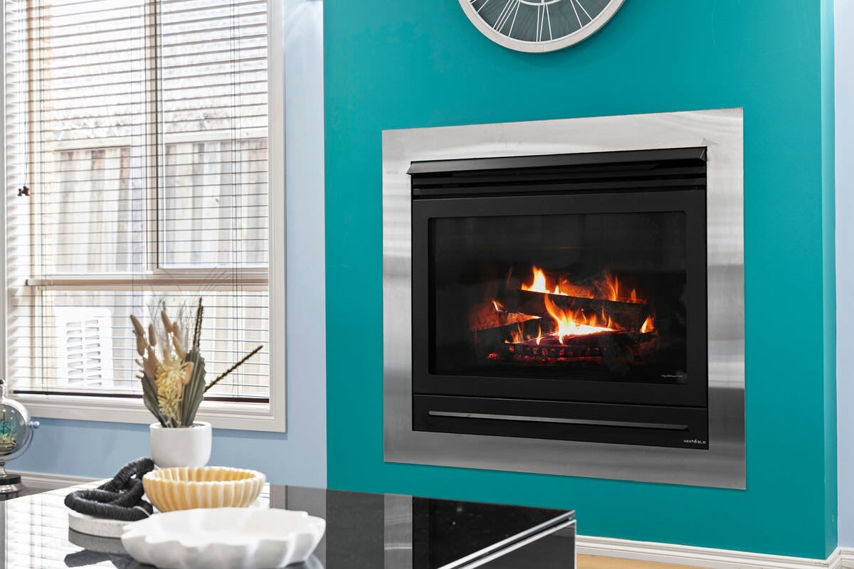 A modern fireplace is set against a vibrant teal wall, featuring a stainless steel surround. A clock hangs above, while natural light filters through the window adorned with blinds. A dark surface with decorative bowls adds a touch of style to the room.