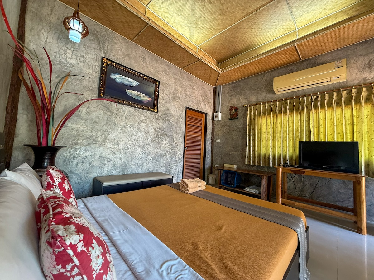 A cozy bedroom showcases a spacious bed adorned with neutral-toned linens and decorative pillows. Natural light filters through sheer yellow curtains, highlighting a wooden desk and a television. A vase with tall, vibrant foliage adds a touch of color to the room's decor.
