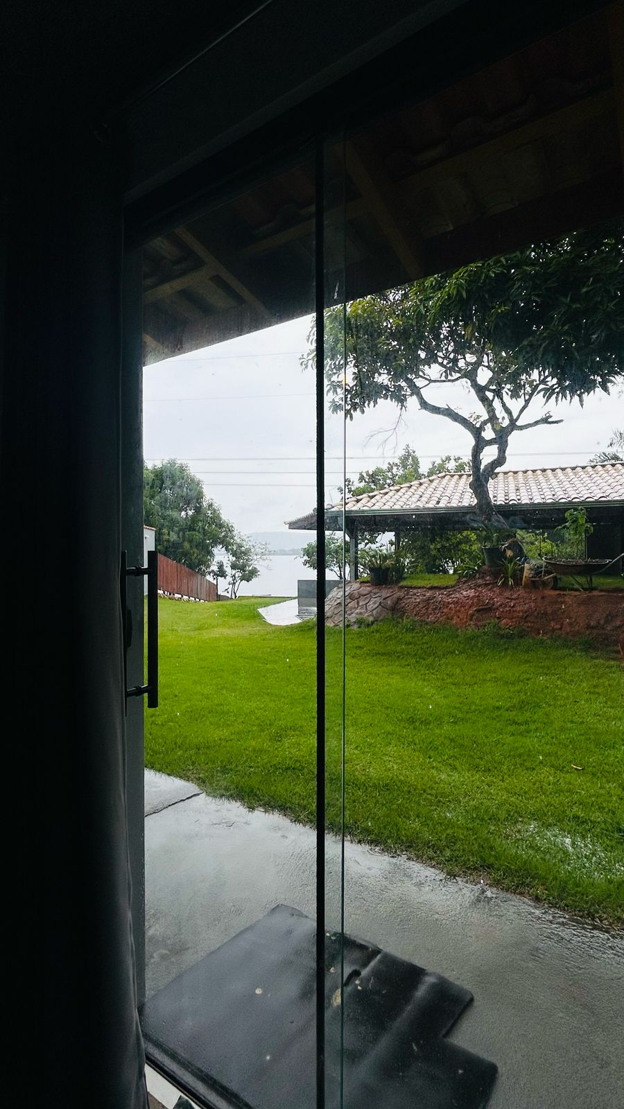 A view from inside shows a glass door reflecting the outdoor scenery. Lush green grass and trees are visible, along with a glimpse of a nearby structure with a tiled roof. The area is illuminated by soft natural light.