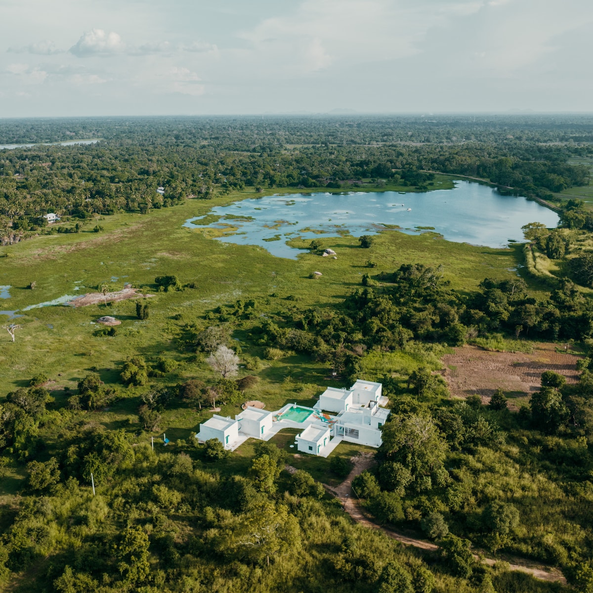 An aerial view captures the villa nestled amidst lush greenery, with a serene lake nearby. The white structure is complemented by a large swimming pool, creating a striking contrast against the surrounding wilderness and expansive landscapes.