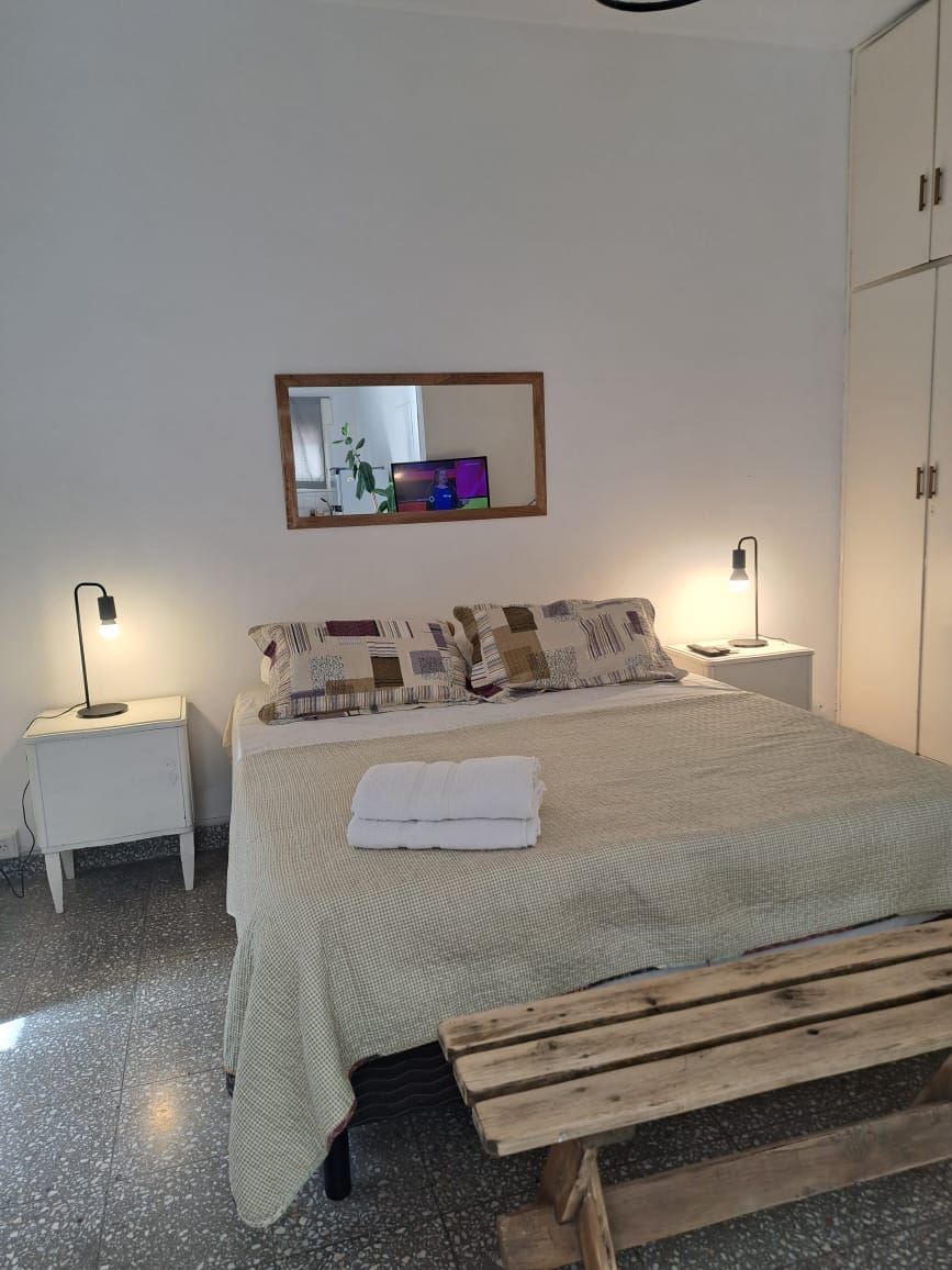 A well-lit sleeping area features a spacious bed with a textured blanket, flanked by two bedside tables each with a lamp. A mirror reflects the room’s light and a plant, while a wooden bench adds a rustic touch to the space.