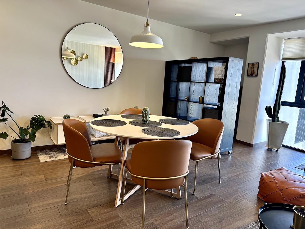 A dining area features a round table surrounded by four brown chairs, complemented by a wall-mounted mirror. A pendant light hangs above the table. Shelves with decorative items are situated in the background, alongside potted plants adding a touch of greenery.