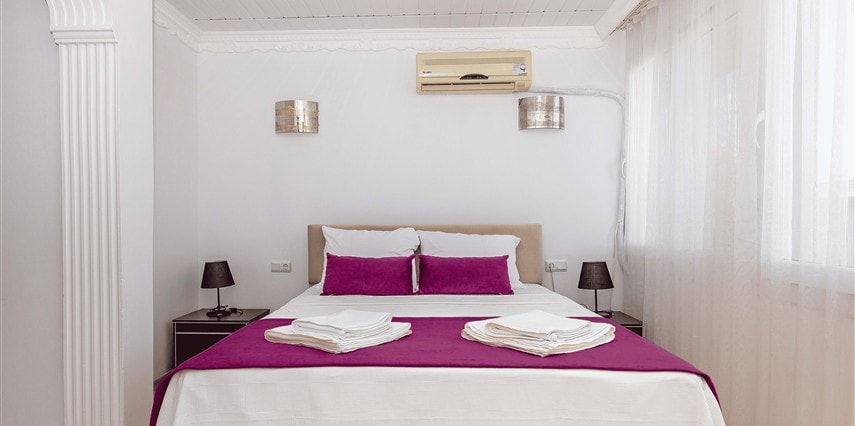A spacious sleeping area features a comfortable bed adorned with white linens and two purple accent pillows. Nightstands with small lamps are placed on either side of the bed. Natural light filters through sheer white curtains, and a wall-mounted air conditioning unit is visible.