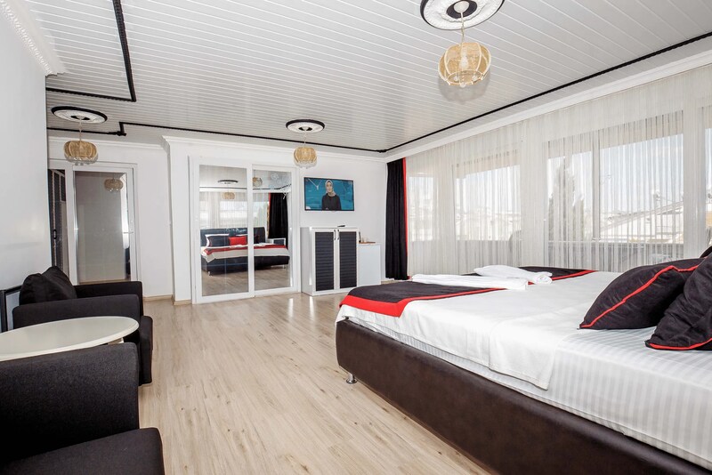 A modern room features a spacious bed with dark pillows and a light blanket. A flat-screen television is mounted on the wall across from the bed. Large windows with sheer curtains allow natural light, and a mirrored wardrobe is positioned beside a seating area with two chairs.