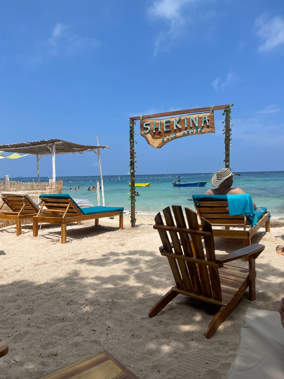 Wooden lounge chairs are arranged on a sandy beach, facing a clear blue sea. A rustic sign reading 'SHEKINAH' is featured prominently, surrounded by lush greenery. A shaded area with a canopy is visible in the background, and small boats can be seen in the distance.