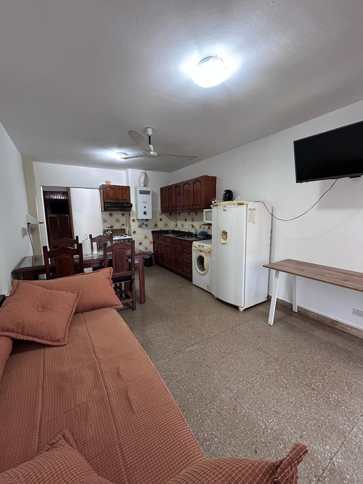 A functional living space includes a cozy sofa, dining area, and kitchen. Brown wooden cabinets house kitchen essentials, while a refrigerator and washing machine are positioned for convenience. Soft lighting from the ceiling illuminates the room, complemented by a wall-mounted television.
