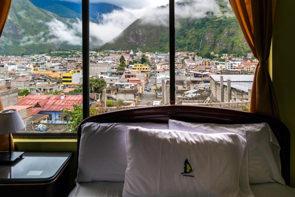 A spacious bed is positioned next to a large window, offering panoramic views of the city and surrounding mountains, partially shrouded in mist. Soft curtains frame the window, allowing natural light to illuminate the room, highlighting the cozy and welcoming atmosphere.