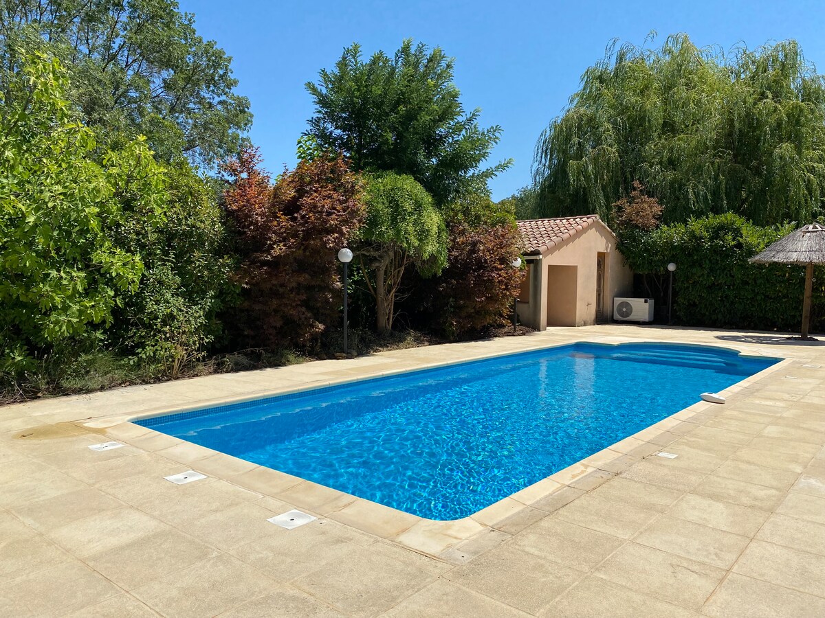A tranquil swimming pool with clear blue water is framed by well-maintained greenery. Surrounding the pool, a spacious paved area offers ample room for relaxation. A small building and a covered area with a thatched roof are visible in the background.