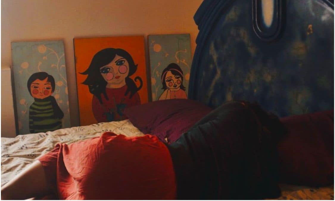 A cozy sleeping area features a large bed with colorful pillows. Behind the bed, three vibrant artworks depicting children are displayed, adding a cheerful touch to the room's atmosphere.