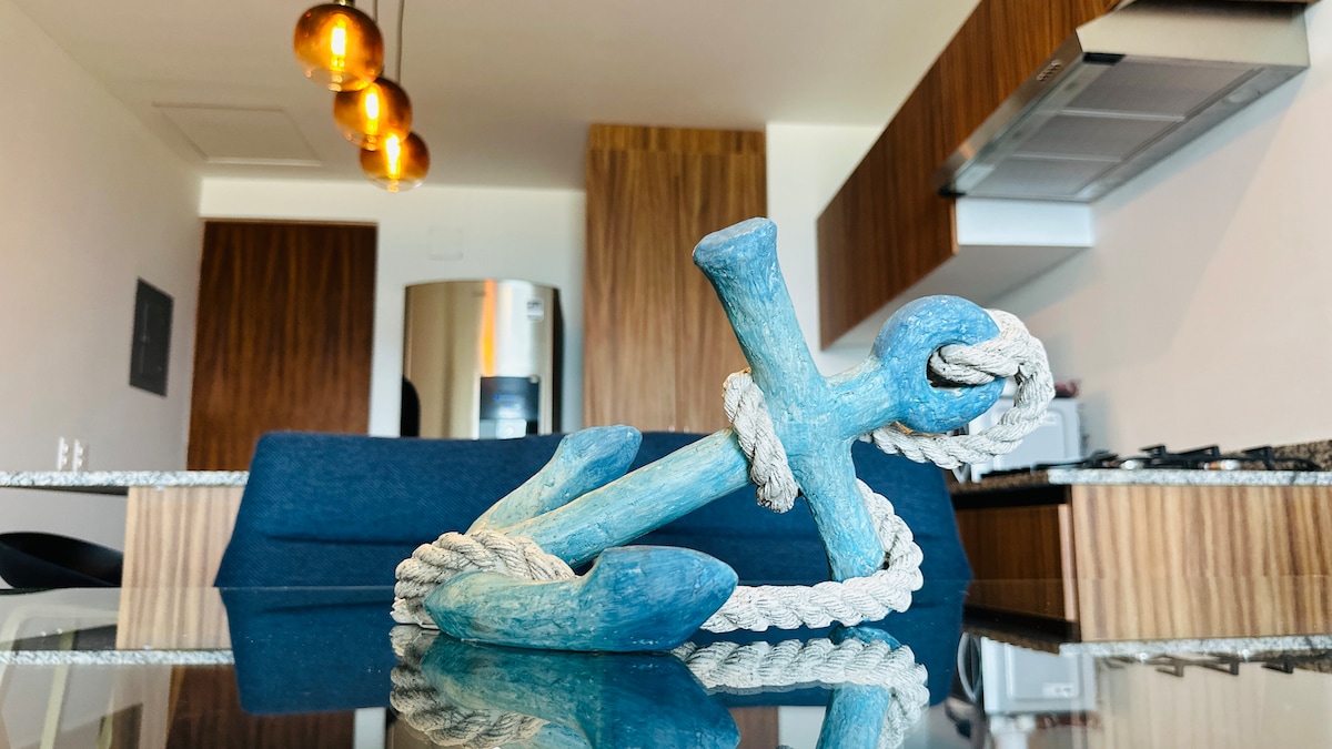 A decorative blue anchor is positioned on a glass surface, complemented by a coiled rope. The background reveals a kitchen area with wooden cabinetry and modern appliances, including a silver refrigerator, creating a nautical-inspired atmosphere.