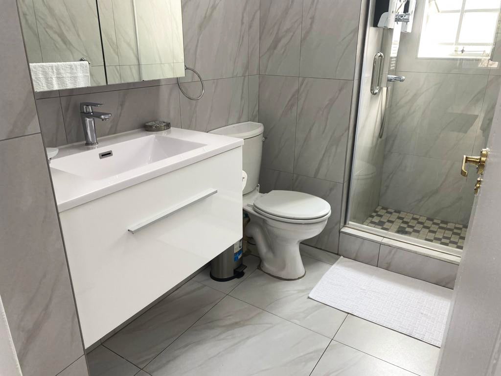 A modern bathroom features light grey tiles and a spacious shower area with glass doors. A sleek white sink cabinet is positioned beneath a large mirror, while a small rug lies on the floor near the toilet. Natural light enters through a window adorned with a sheer covering.