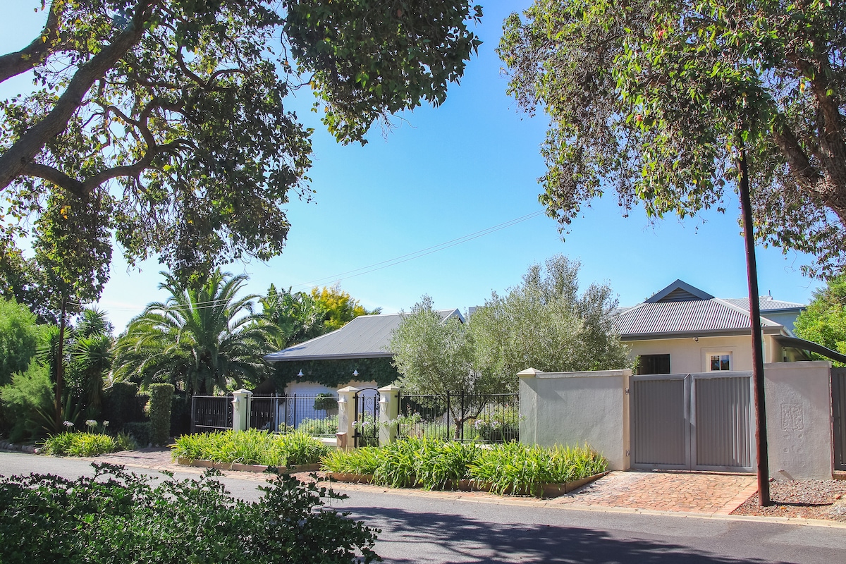 A welcoming exterior is seen, featuring a well-maintained garden with green foliage and palm trees. The main entrance is marked by a gate, with a path leading to the house, which is set against a clear blue sky.