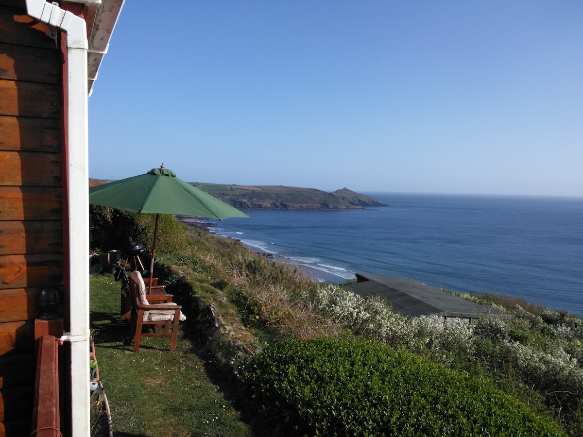 A serene coastal view is presented, featuring a large green umbrella shading a comfortable seating area. The lush garden leads down to the waves lapping against the shore, with a clear blue sky above and distant cliffs visible on the horizon.