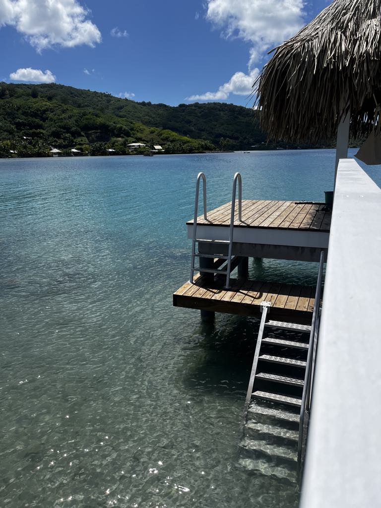 A wooden deck extends over clear turquoise waters, featuring a ladder for easy access to the lagoon. Lush green hills rise in the background, framed by a bright sky with scattered clouds. A thatched roof provides shade at the edge of the deck.