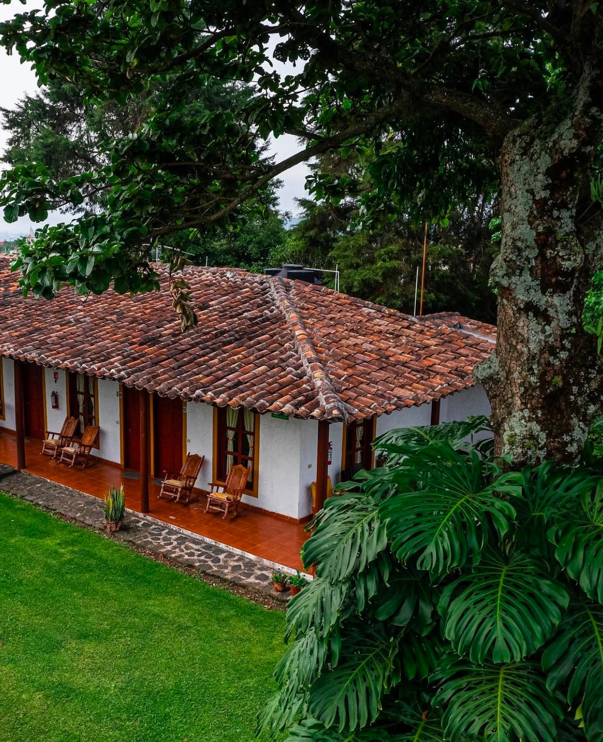 A tranquil view of a traditional house is framed by lush greenery. The structure features a tiled roof and wooden rocking chairs on a covered porch. A well-maintained garden with grass and foliage surrounds the house.