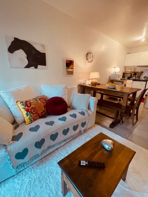 A light and inviting living space is highlighted by a comfortable white sofa draped with a heart-patterned throw. A wooden dining table is set nearby, accompanied by rustic chairs, while kitchen elements are visible in the background. A wall clock and artwork add character to the area.