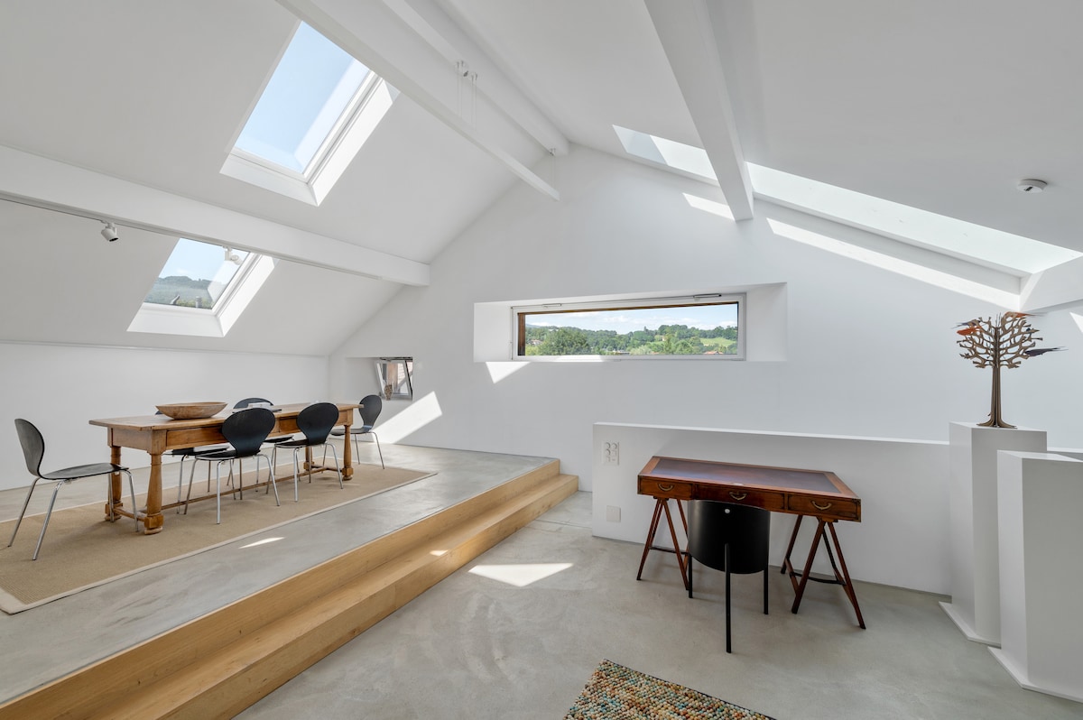 A bright and airy space features a wooden dining table surrounded by six chairs. Large skylights illuminate the room, enhancing the expansive feeling. A wooden desk is placed adjacent to a small decorative tree, with views of the surrounding mountains visible through the window.