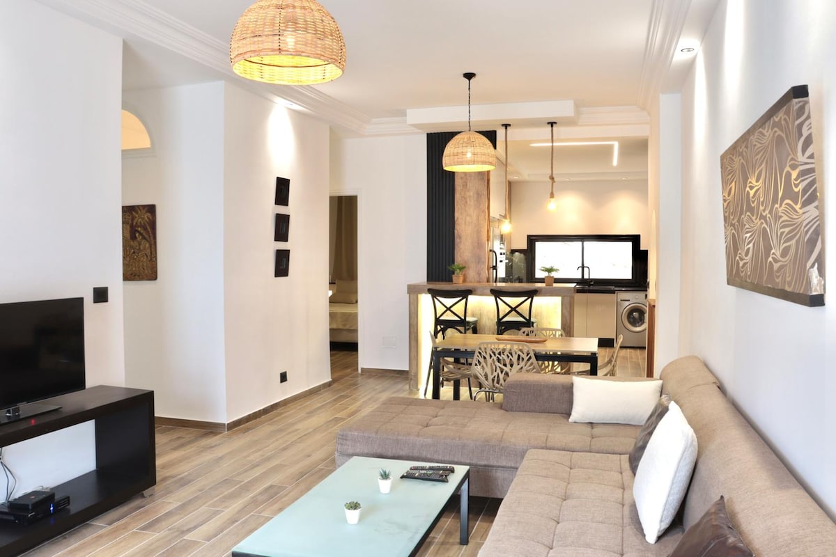 A modern living area features a comfortable sectional sofa arranged around a sleek glass coffee table. Soft lighting is provided by woven pendant lamps, enhancing the clean lines of the room. In the background, an open kitchen area displays contemporary appliances.