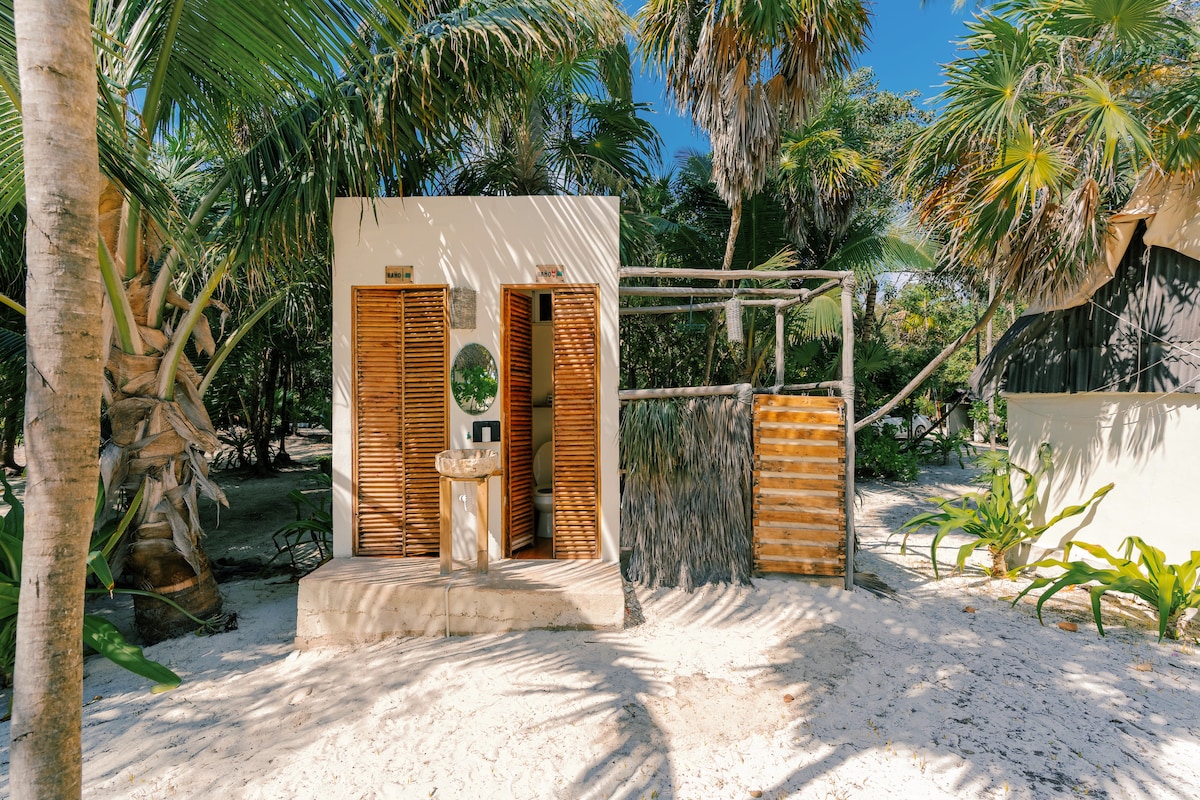 A cabin entrance is framed by lush tropical foliage, showcasing wooden slatted doors and a round mirror by the doorway. Natural materials are incorporated into the design, with a rustic wooden fence creating privacy. The sandy pathway leads to a serene environment.