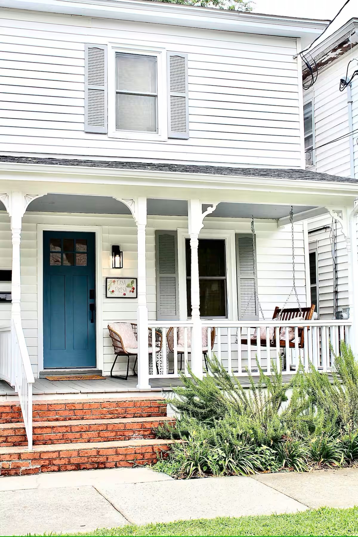 The front porch of the duplex features a light blue door framed by white railings. A wooden swing is positioned beside a small table and two chairs. Lush green plants and brick steps lead to the entrance, adding a welcoming touch.