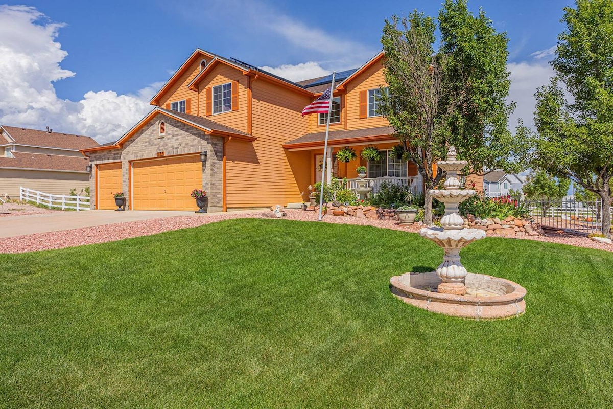 The exterior of a custom-built property is presented, featuring a vibrant orange façade and stone accents. Lush green grass surrounds the home, with mature trees and flower beds adding to the landscape. A decorative fountain is centrally placed in a circular stone area, and an American flag is displayed.