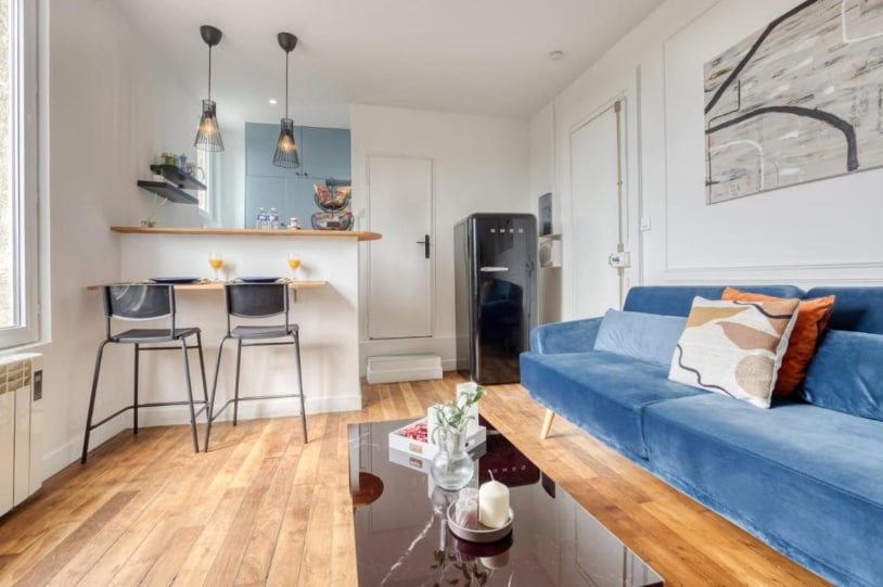 A bright living area features a blue sofa adorned with decorative cushions. Noticeable are the wooden floors and a glass coffee table at the center. A small dining area with two black chairs sits adjacent to a kitchenette equipped with modern appliances, including a black refrigerator.