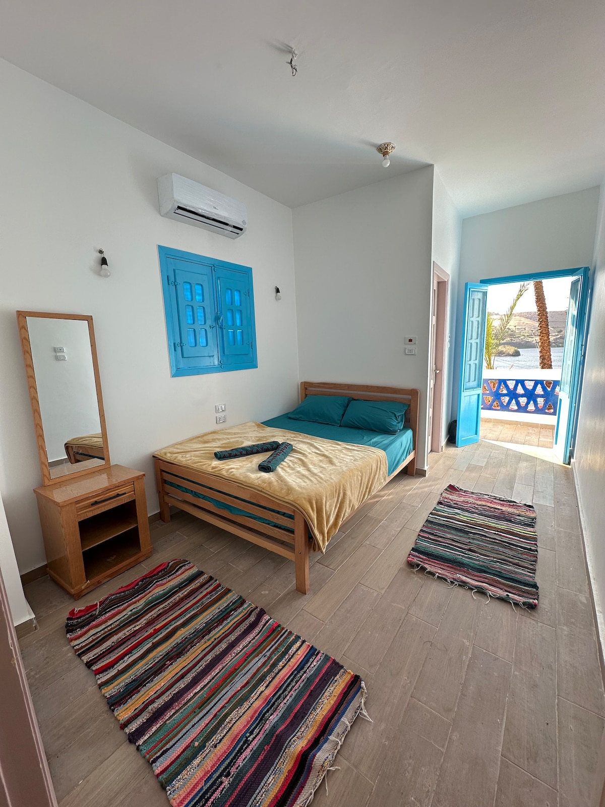 A bright room features a wooden bed with soft bedding and two colorful area rugs on a tiled floor. A mirror stands on a small dresser. Blue doors frame a view of the outdoor area, allowing natural light to fill the space.
