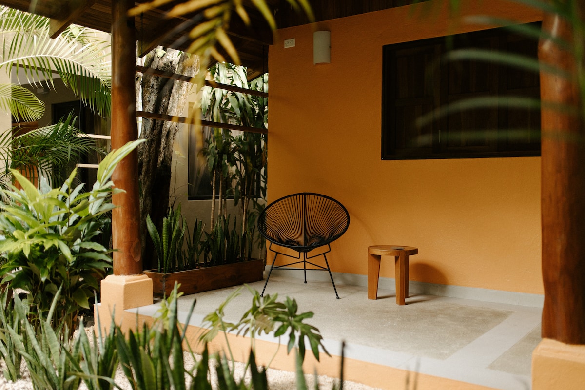 An outdoor seating area is situated beneath a wooden porch, featuring a black acapulco chair and a small wooden table. Lush greenery surrounds the space, providing a serene atmosphere. The walls are painted in a warm orange hue, complementing the natural surroundings.