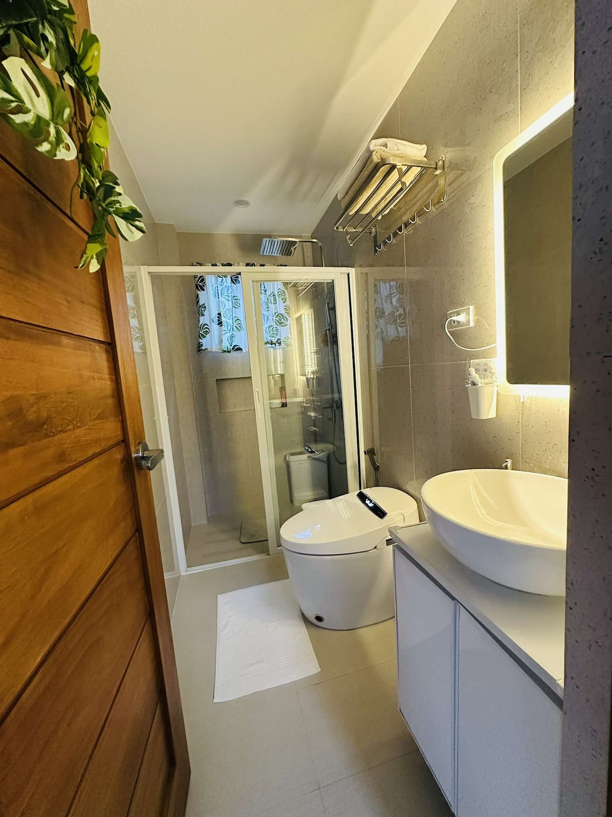 A modern bathroom is depicted, featuring a glass-enclosed shower area with floral-patterned curtains. A sleek, circular sink is positioned on a minimalist vanity, complemented by a smart toilet. Soft lighting enhances the clean, neutral tones of the tiled walls and floor.