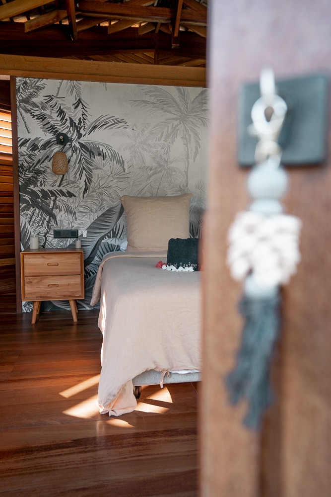 The bedroom features light wood flooring and is decorated with a tropical leaf mural on one wall. A spacious bed with neutral linens is positioned beside a wooden nightstand. Soft natural light filters through wooden slats, creating a relaxed ambiance.