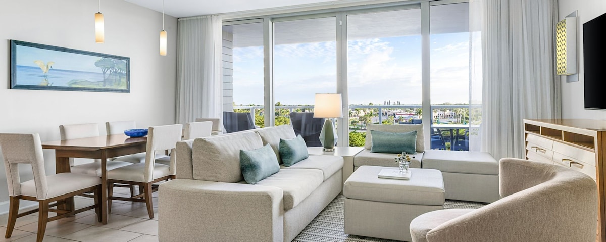 A light-filled living area features a soft sofa and an armchair, complemented by decorative cushions. A dining table with light-colored chairs is visible in the background, and floor-to-ceiling windows offer expansive views of the outdoor scenery beyond.