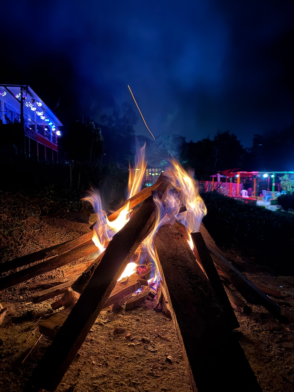 A vibrant campfire is captured, with flames dancing above a stack of wooden logs. The background reveals an illuminated outdoor space with colorful lights, hinting at social areas nearby. Dark foliage frames the scene, contributing to the evening ambiance.