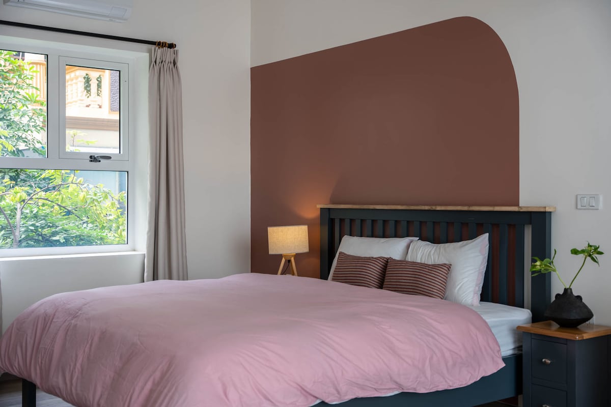 A spacious bed with a soft pink blanket is positioned against a two-tone wall featuring a warm brown arch. The area is complemented by a bedside table with a plant and lamp, while natural light from the window illuminates the room.