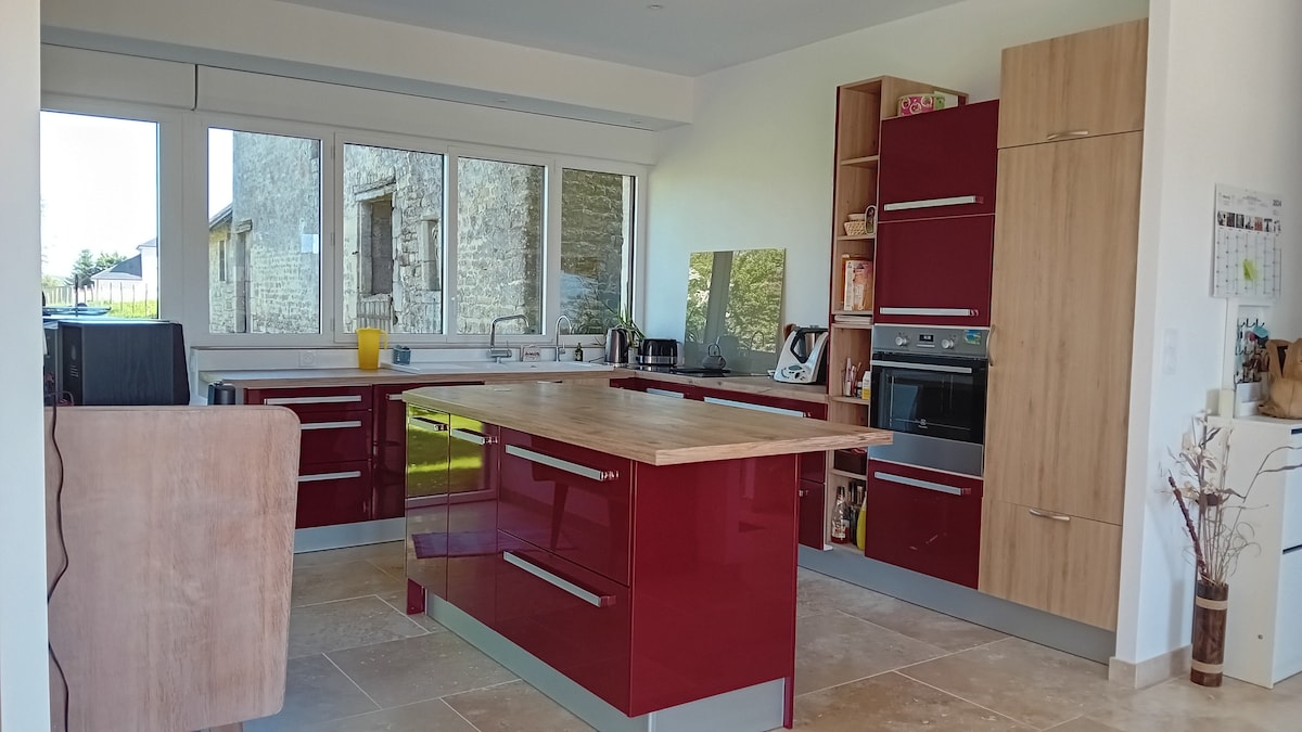 A modern kitchen features glossy red cabinetry and a large wooden island at its center. Natural light floods in through expansive windows, illuminating stainless steel appliances and a cozy seating area. A casual dining space is visible, creating a functional and inviting atmosphere.