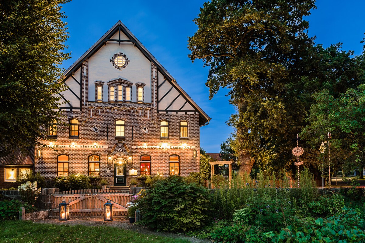 The historic country house features ornate brickwork and a distinct architecture, highlighted by warm lighting that outlines the windows and doors. Lush greenery frames the entrance, leading to a charming patio area. A serene evening sky provides a tranquil backdrop to the inviting exterior.