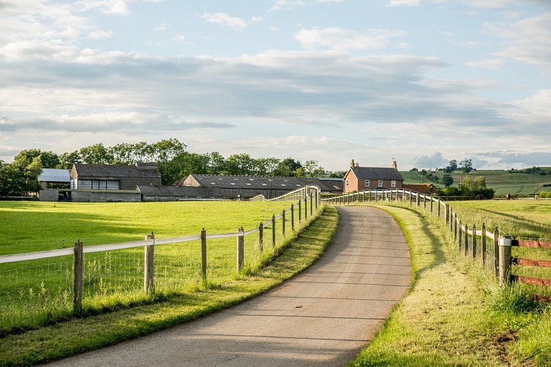 A winding path leads through lush green fields toward a cluster of farm buildings. The landscape is complemented by a wooden fence lining the road, and gentle hills rise in the background beneath a partly cloudy sky.