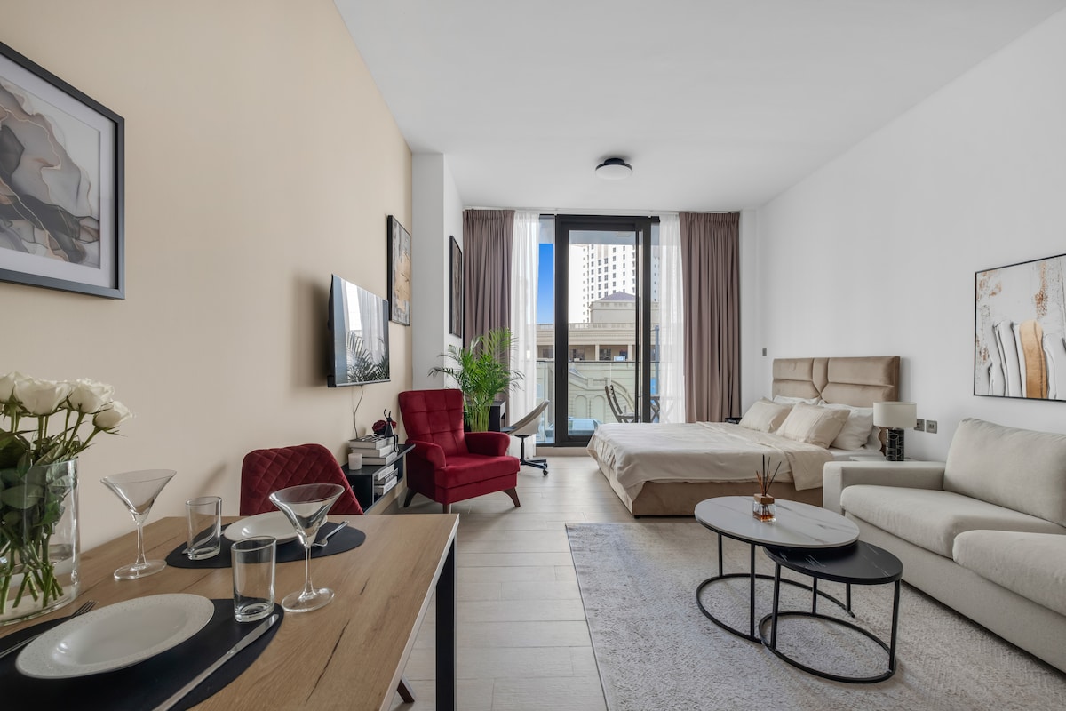 A modern studio layout features a comfortable bed, a sofa, and a dining table set for two. Light-colored walls complement the contemporary furnishings. Large windows provide natural light and an inviting view, while potted greenery adds a touch of vibrancy to the space.