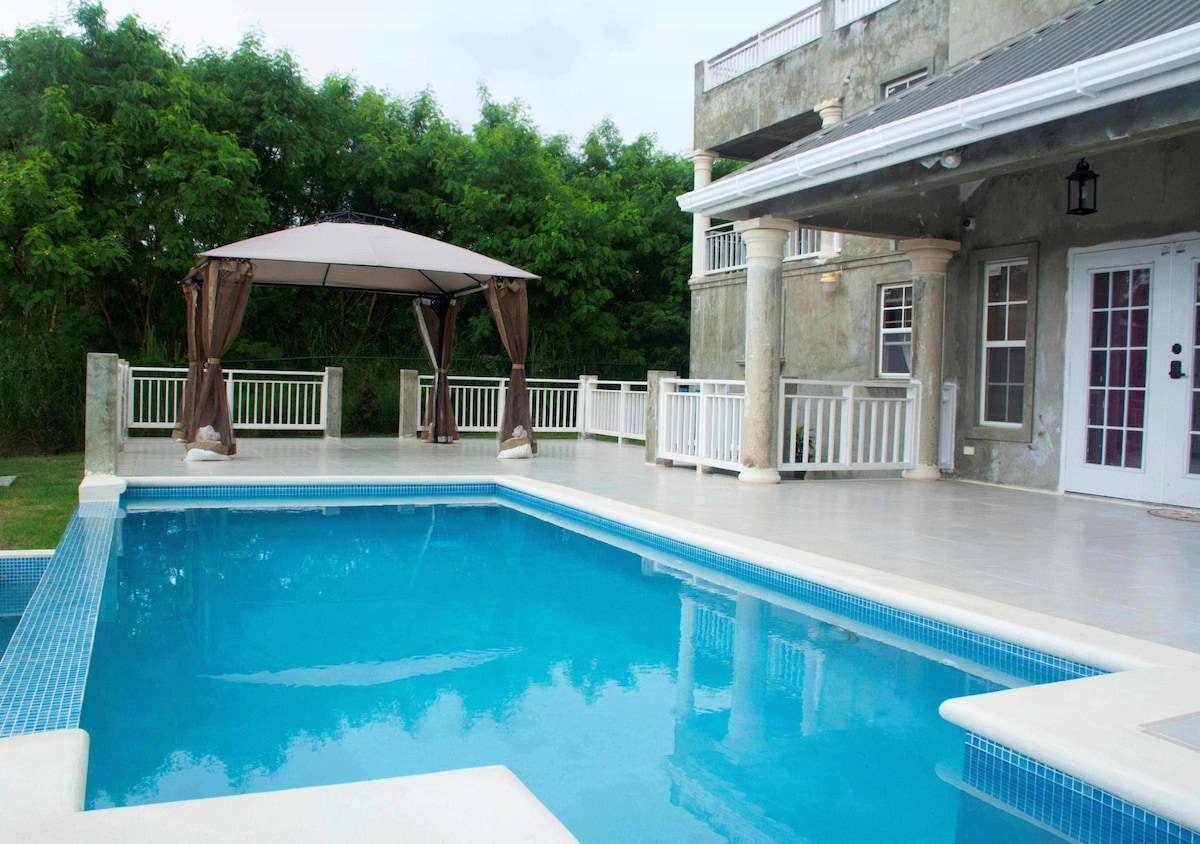 An inviting infinity pool is highlighted, featuring clear blue water surrounded by a spacious deck. A shaded gazebo stands nearby, bordered by lush greenery. The pool area offers an ideal setting for relaxation and gatherings, with ample space for outdoor enjoyment.
