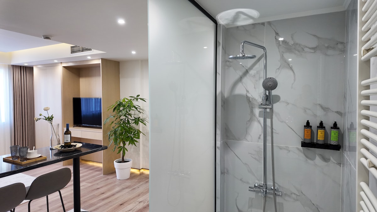 A modern bathroom area is displayed, featuring a glass-enclosed shower with a sleek rain showerhead. Nearby, a small dining space is visible, showcasing a table set for two with a bottle of wine and glasses. Natural light highlights the minimalistic design and soothing color scheme.