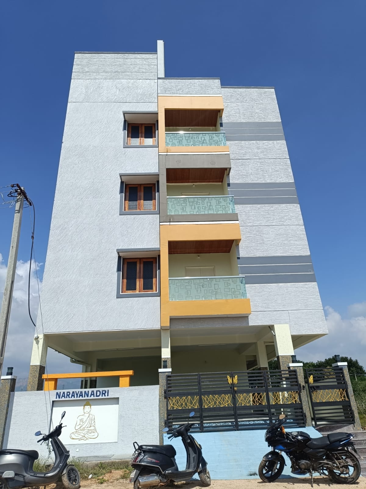 A modern three-story building features a combination of light and dark gray exterior walls. Balconies with glass railings are visible on each floor. The entrance is marked by signage, and parked scooters can be seen in the foreground against a clear blue sky.