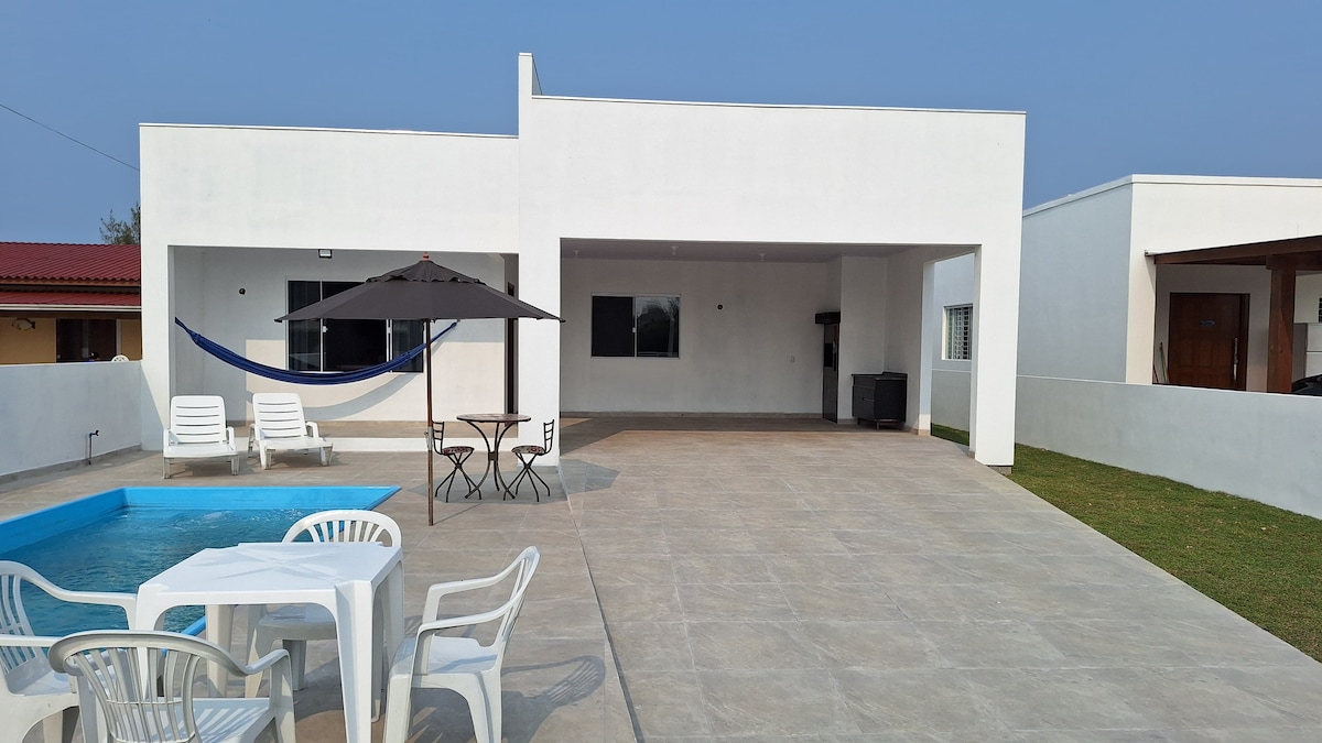 An outdoor area features a private pool surrounded by lounge chairs. A shaded dining table with plastic chairs is present, along with a hammock hanging in the background. The clean, modern architecture is complemented by a spacious tiled patio and manicured grass.