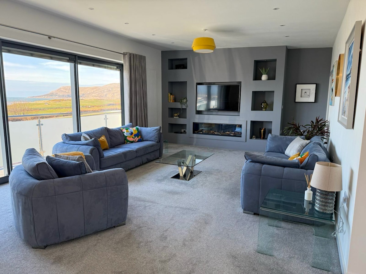 A spacious living area is shown, featuring large sliding glass doors that offer panoramic sea views. Two comfortable gray sofas are positioned around a modern glass coffee table, with subtle decorative touches like colorful cushions. A wall-mounted television and shelving complete the inviting space.