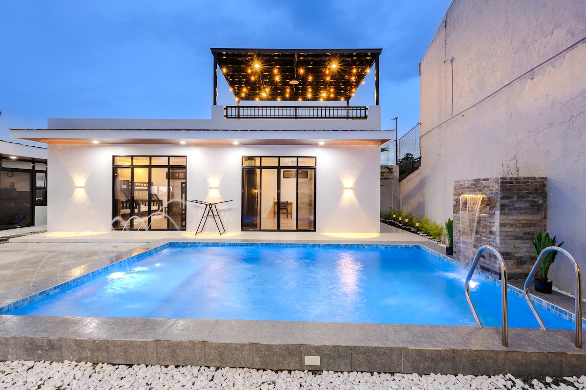 A modern villa is illuminated under the evening sky, featuring an inviting pool with clear blue water. Its sleek design includes large windows and a spacious outdoor deck adorned with soft lighting. A cascading fountain adds a tranquil element to the surrounding area.